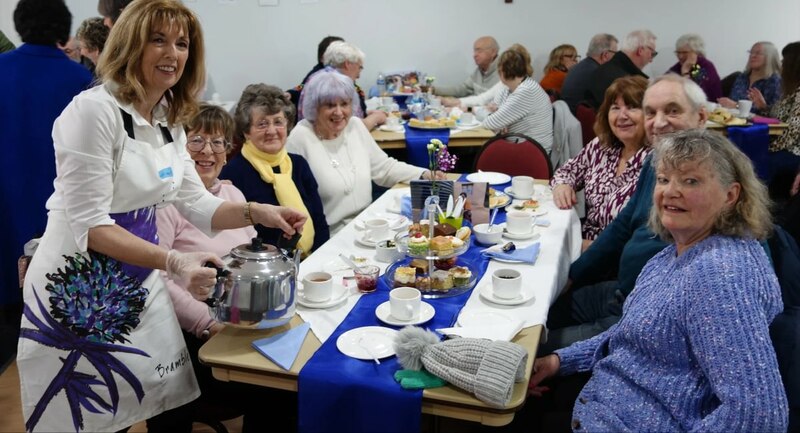 A table of people smiling with one person standing up holding a teapot, looking at the camera